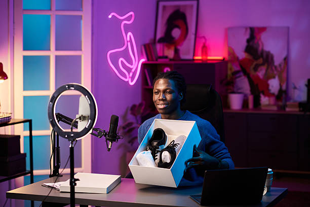 Black person unboxing sneakers in modern workspace with bright neon lights. Studio decorated with various artworks and gadgets creating a trendy and vibrant environment